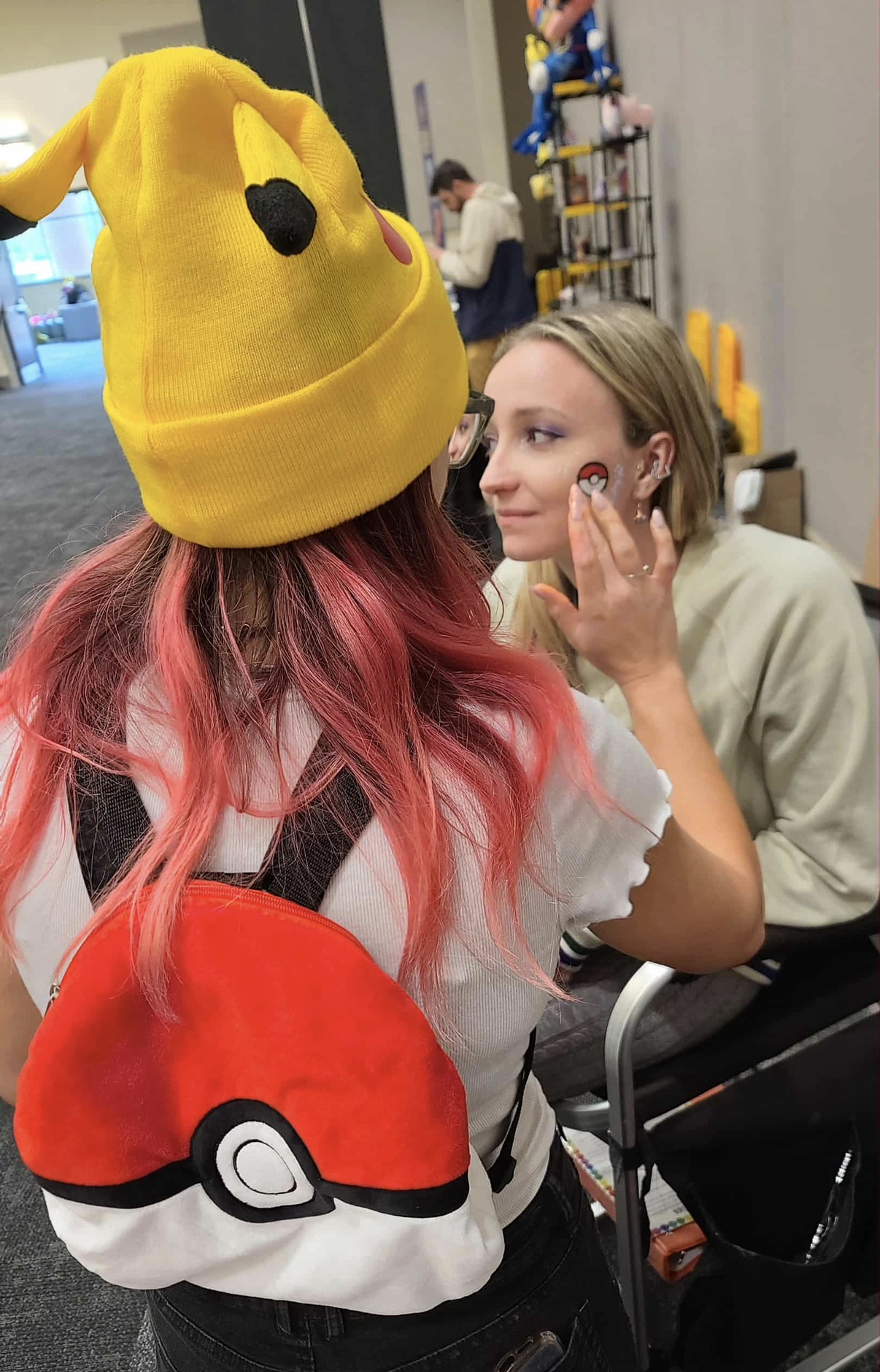 Sadie in a Pikachu hat painting a Pokeball on someone's face at a convention